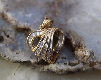 May include: A gold heart-shaped pendant with a series of baguette-cut diamonds. The pendant has a curved design, with the diamonds set along the curves. The bail is a simple loop. The pendant is displayed on a light blue and gray stone surface.