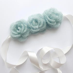 Puede incluir: Una delicada diadema con tres flores de ganchillo de color azul claro y esponjosas. Las flores están unidas a una cinta de satén blanco, que está rizada y drapeada sobre un fondo blanco. Las flores son suaves y texturizadas.