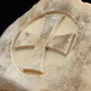 May include: A carved stone featuring a cross design within a circular frame. The stone has a natural, textured appearance with variations in color, ranging from beige to light brown. The cross is deeply etched into the stone.