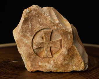 May include: A carved stone with a cross design. The stone is a light brown color and has a rough texture.
