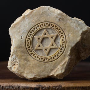 Może przedstawiać: Beżowy kamień z rzeźbioną Gwiazdą Dawida w kółku. Kamień jest szorstki i fakturowany.