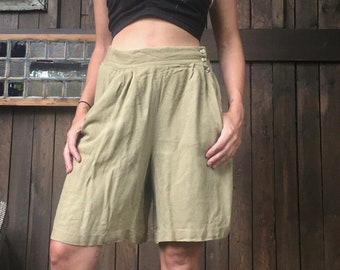 May include: A woman wearing a black bandeau top and khaki linen shorts. She is standing in front of a wooden wall with a large antler mounted on it. She is wearing a black hat and has long blonde hair.