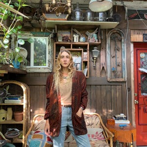 May include: A woman wearing a brown patterned jacket over a light-colored top and blue jeans. The jacket has a dark brown zebra-like pattern. She is standing in front of a wooden wall with shelves and decorative items.