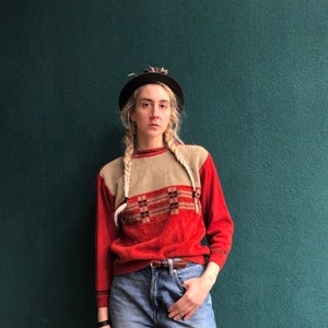 May include: A woman wearing a red and beige sweater with a geometric pattern, a black hat, and blue jeans.