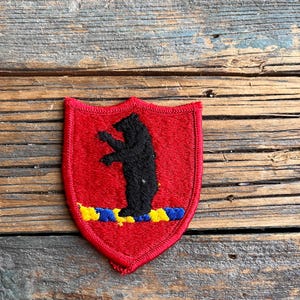 May include: A red embroidered patch featuring a black bear standing on a blue and yellow stripe. The patch is shaped like a shield.