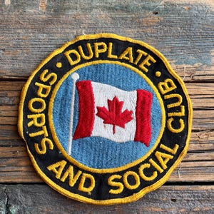 May include: A black and yellow embroidered patch with the text "Duplate Sports and Social Club" in a circle. A Canadian flag is in the center of the circle.