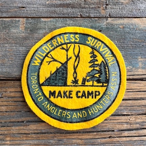May include: A yellow felt patch with green text and a black and green illustration of a campsite. The text reads "Wilderness Survival Toronto Anglers and Hunters Assn. Make Camp".