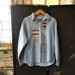 May include: A light blue long-sleeved button-down shirt with a geometric embroidery design in brown, white, and grey. The shirt has a collar and a button closure.