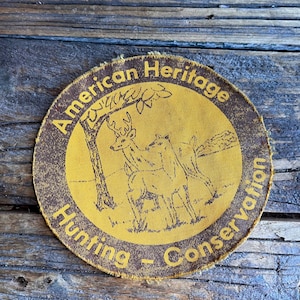 May include: A yellow and brown round patch with the text "American Heritage Hunting - Conservation" and a drawing of a deer and a bear in a woodland setting.