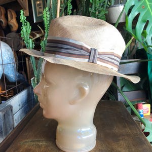 May include: A tan straw hat with a brown and white striped band. The hat is sitting on a mannequin head.