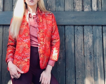 Può includere: Una donna indossa una giacca trapuntata rossa con dettagli dorati e pantaloni bordeaux. È in piedi davanti a un muro di legno e indossa occhiali da sole gialli.