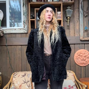 May include: A woman wearing a black textured coat and a black hat. The coat is open, revealing a light-coloured turtleneck. She is standing in front of a wooden wall with a window and decorative items.