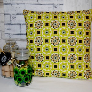 Puede incluir: Una almohada de tiro verde y marr&oacute;n con un dise&ntilde;o geom&eacute;trico. La almohada est&aacute; sentada sobre una superficie blanca frente a dos frascos de vidrio llenos de botones.