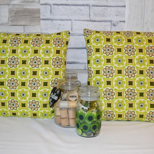 Puede incluir: Dos almohadas de tiro con estampado verde y marr&oacute;n sobre fondo blanco. Las almohadas est&aacute;n sobre una superficie blanca con dos frascos de vidrio llenos de botones y carretes de hilo.