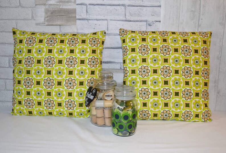 Puede incluir: Dos almohadas de tiro con estampado verde y marr&oacute;n sobre fondo blanco. Las almohadas est&aacute;n sobre una superficie blanca con dos frascos de vidrio llenos de botones y carretes de hilo.