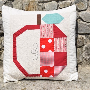 May include: Square white cushion with a patchwork apple design. The apple features red and white polka dot, gingham, and floral patterns. The stem is brown, and the leaf is a teal blue geometric pattern. The apple is outlined in red.