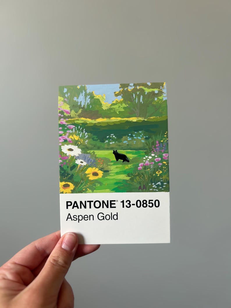 K&ouml;nnte beinhalten: Eine rechteckige Karte mit einem Gartenbild, einschlie&szlig;lich einer schwarzen Katze. Die Karte zeigt den Text "PANTONE 13-0850 Aspen Gold". Die Gartenszene zeigt Blumen, B&auml;ume und einen blauen Himmel.
