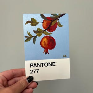 Könnte beinhalten: Gemälde von Granatäpfeln an einem Ast vor hellblauem Hintergrund. Das Kunstwerk befindet sich auf einem Pantone 277 Farbfeld. Die Granatäpfel sind rot und orange, mit grünen Blättern. Die Initialen des Künstlers sind oben rechts.