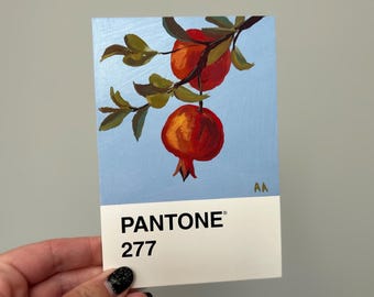 May include: A painting of pomegranates on a branch against a light blue background. The artwork is on a Pantone 277 color swatch. The pomegranates are red and orange, with green leaves. The artist's initials are in the upper right corner.