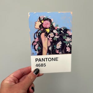 Könnte beinhalten: Kleines Kunstwerk mit einer Frau mit langen schwarzen Haaren, die mit rosa und weißen Blumen geschmückt sind. Das Kunstwerk befindet sich auf einem weißen Pantone-Farbfächer mit der Aufschrift "PANTONE 4685". Das Profil der Frau ist vor einem hellblauen Hintergrund zu sehen.