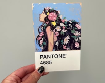 May include: A small artwork featuring a woman with long black hair adorned with pink and white flowers. The artwork is on a white Pantone color swatch labeled "PANTONE 4685". The woman's profile is visible against a light blue background.