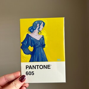 May include: A rectangular art print featuring a woman in a blue dress against a yellow background. The print is labeled "PANTONE 605" in white text at the bottom. The artwork is held by a hand with a dark red nail polish.