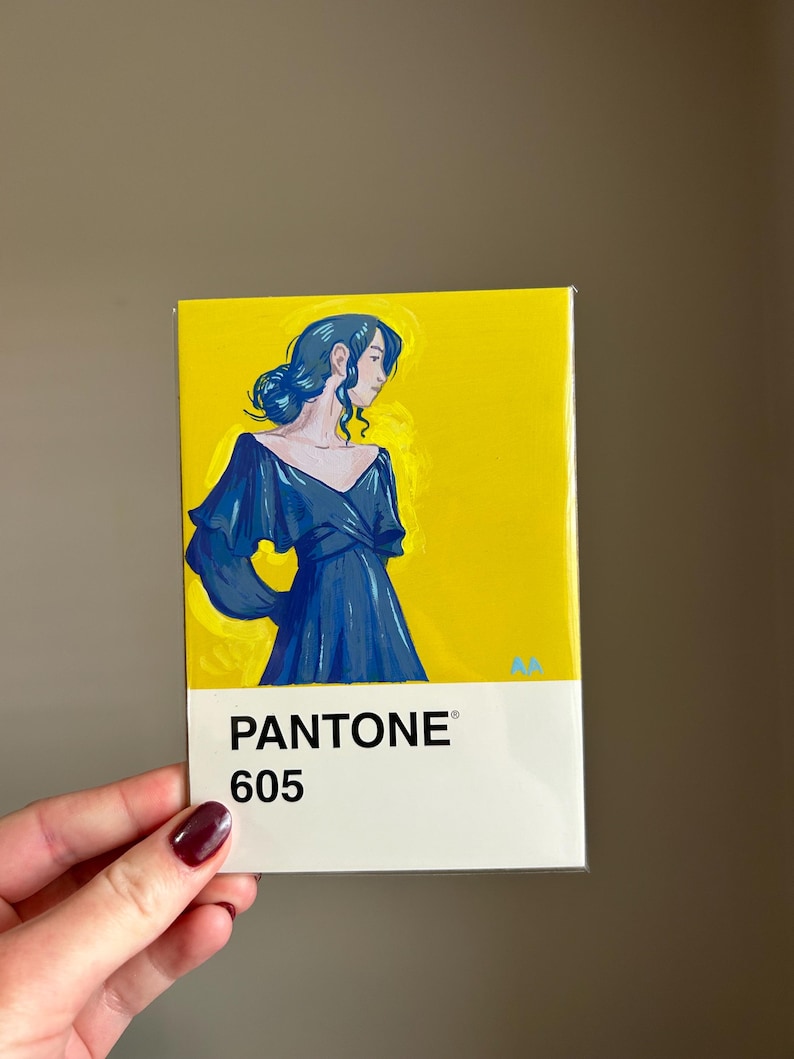 May include: A rectangular art print featuring a woman in a blue dress against a yellow background. The print is labeled "PANTONE 605" in white text at the bottom. The artwork is held by a hand with a dark red nail polish.