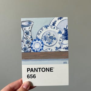 May include: A Pantone color swatch card featuring a still life painting of blue and white ceramic plates and a vase. The card displays the color code "656". The artwork depicts floral patterns and a windmill.