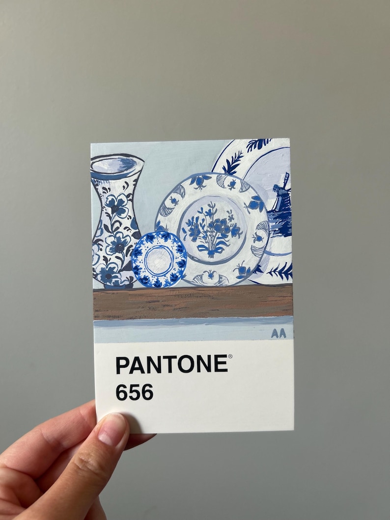 May include: A Pantone color swatch card featuring a still life painting of blue and white ceramic plates and a vase. The card displays the color code "656". The artwork depicts floral patterns and a windmill.