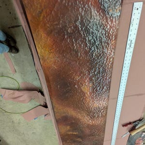 May include: A rectangular sheet of hammered copper with a variegated patina featuring shades of deep brown, orange, and metallic red. A white metal ruler is positioned along the right edge, showing a length of approximately 28 inches.