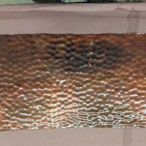 May include: A rectangular copper sheet featuring a hammered, dimpled texture across its entire surface. The metal displays a gradient of warm brown and reddish-orange tones, with reflective highlights catching the light across the uneven, textured finish.