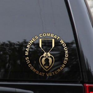 May include: A gold-coloured decal featuring the words "US Marines Combat Wounded Combat Veteran" encircling a Purple Heart medal. The medal has a heart-shaped centre with two silhouettes.