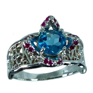 May include: A silver ring featuring a large, round, light blue gemstone at the center, surrounded by smaller red gemstones. The ring has an ornate, openwork design on the band.