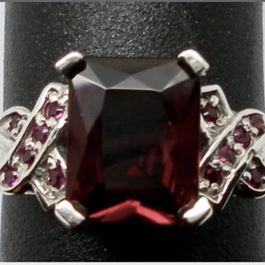 May include: A silver ring with a large, dark red, rectangular gemstone surrounded by smaller, red gemstones.