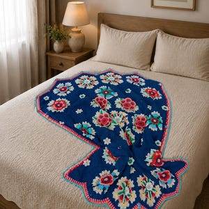 Può includere: Una coperta blu navy con motivo floreale e bordo smerlato, esposta su un letto. La coperta presenta fiori rossi, bianchi e gialli. Il bordo è rosa e turchese. Il letto ha lenzuola beige.