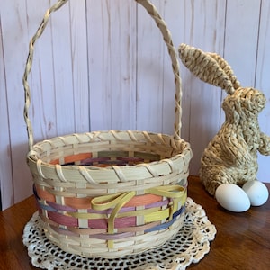 K&ouml;nnte beinhalten: Ein geflochtener Weidenkorb f&uuml;r Ostern mit einer bunten Schleife. Der Korb steht auf einer wei&szlig;en H&auml;keldeckchen auf einer Holzoberfl&auml;che. Im Hintergrund sind zwei wei&szlig;e Eier und ein geflochtener Strohhase zu sehen.