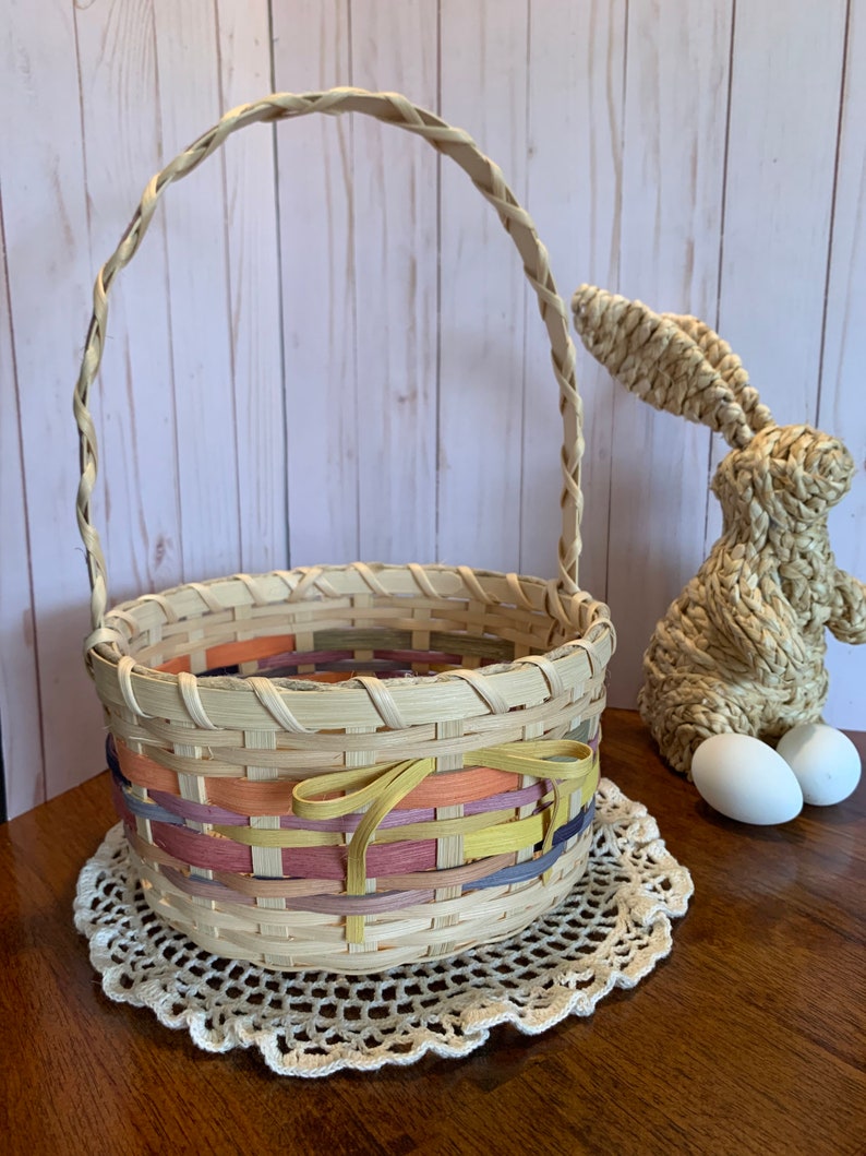 K&ouml;nnte beinhalten: Ein geflochtener Weidenkorb f&uuml;r Ostern mit einer bunten Schleife. Der Korb steht auf einer wei&szlig;en H&auml;keldeckchen auf einer Holzoberfl&auml;che. Im Hintergrund sind zwei wei&szlig;e Eier und ein geflochtener Strohhase zu sehen.