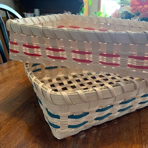 May include: Two rectangular woven baskets made of light-colored splint material. The top basket features horizontal bands of bright red, while the bottom basket features horizontal bands of teal blue, both woven into the light-colored structure.