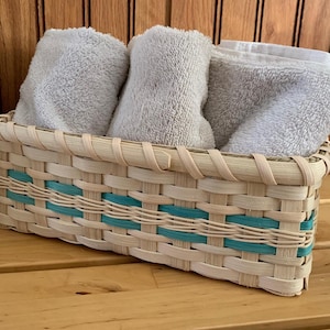 May include: A rectangular woven basket with light tan natural reeds and a horizontal teal accent band. Three folded light gray terry cloth towels are neatly arranged inside the basket, filling the interior space with their soft, textured fabric.