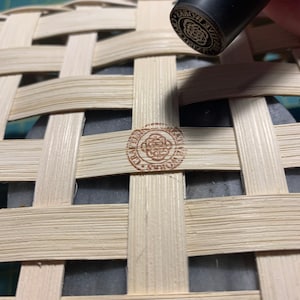 K&ouml;nnte beinhalten: Nahaufnahme eines braunen Holzkorbes mit geflochtenem Muster. Ein Stempel mit dem Text "WORLD'S BEST" wird in den Korb gedr&uuml;ckt.