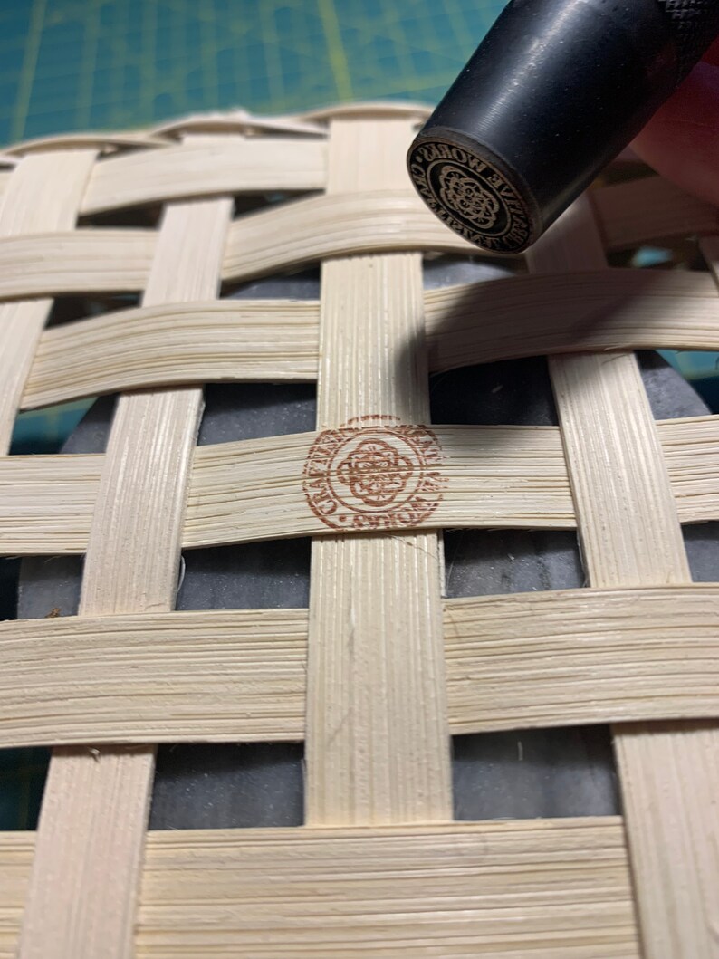 K&ouml;nnte beinhalten: Nahaufnahme eines braunen Holzkorbes mit geflochtenem Muster. Ein Stempel mit dem Text "WORLD'S BEST" wird in den Korb gedr&uuml;ckt.