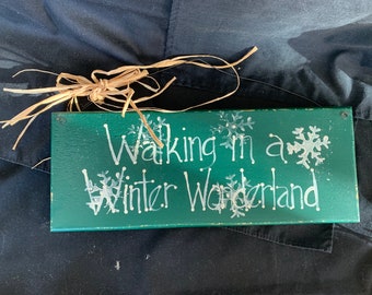 May include: A green wooden sign with white painted text that reads "Walking in a Winter Wonderland". The sign has a rustic look and is decorated with white snowflake designs.