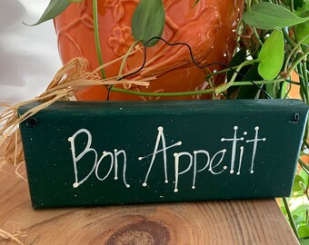 May include: A green wooden sign with white lettering that reads "Bon Appetit". The sign is hanging from a twine string and is sitting on a wooden surface.