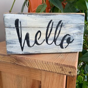 Peut inclure: Une petite boîte en bois avec une finition blanchie à la chaux et un lettrage noir qui dit "hello".
