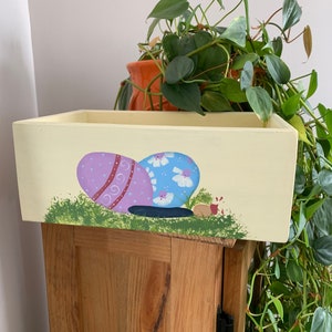 Könnte beinhalten: Ein gelber Holz-Pflanzkasten mit einem bemalten Design aus zwei Ostereiern und einem kleinen roten Marienkäfer. Die Eier sind mit lila, blau und weißer Farbe verziert.