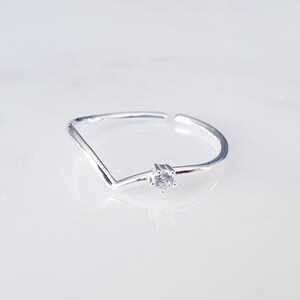 Może przedstawiać: Srebrny pierścionek z małym, przezroczystym kamieniem szlachetnym osadzonym w środku. Pierścionek ma kształt prostego, otwartego kwadratu.