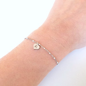Könnte beinhalten: Ein silbernes Kettenarmband mit einem kleinen Herz-Anhänger, der mit dem Buchstaben "K" graviert ist.