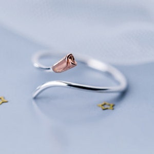 May include: A silver ring with a rose-gold rosebud design. The ring is open and adjustable.