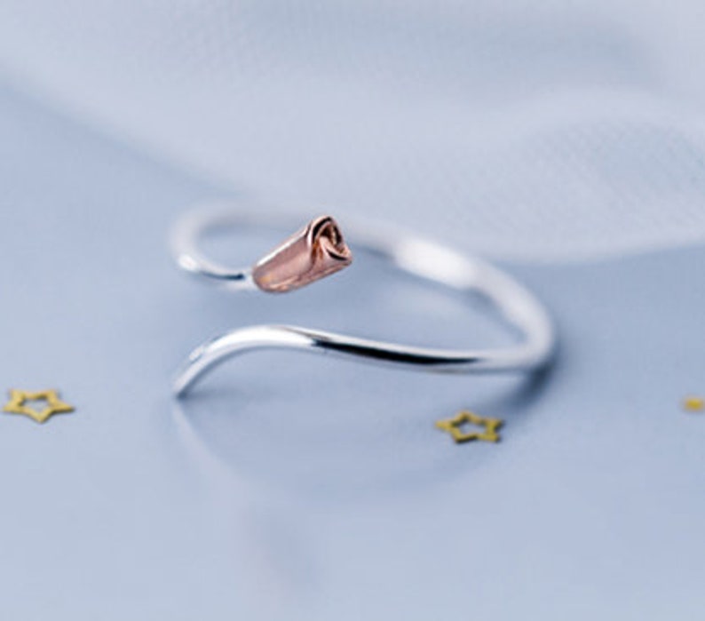 May include: A silver ring with a rose-gold rosebud design. The ring is open and adjustable.