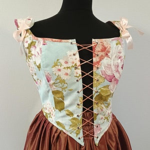 May include: A light blue corset with a floral print and a brown satin skirt. The corset has a lace-up front and is tied with a pink ribbon at the shoulders.